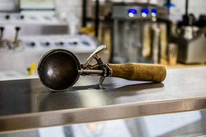 Vintage metal ice cream scoop with wooden handle resting on stainless steel counter in commercial kitchen.