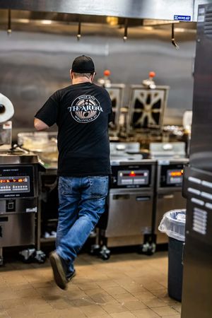 A cook in a black The Arena t-shirt and jeans works in a commercial kitchen with fryers and stainless steel equipment.