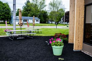 Outdoor patio area with picnic tables, black mulch, pink flowers in green pot, and green lawn with houses in background.