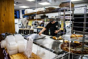 Worker prepares food behind sneeze guard at busy pizza restaurant counter with metal shelving and fresh pizzas.