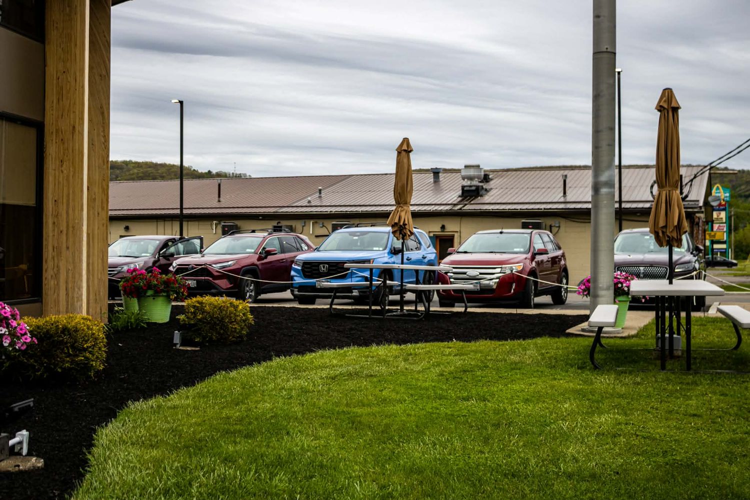Outdoor parking lot with colorful cars, patio umbrellas, and green lawn near a commercial building under cloudy skies.