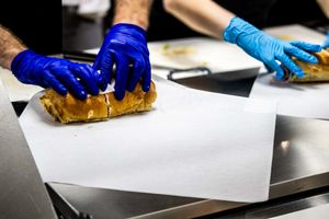 Workers in blue gloves preparing a sandwich wrap on white parchment paper in a commercial kitchen.