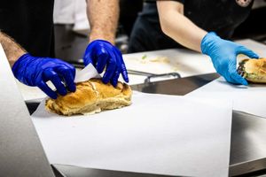 Chef wearing blue gloves preparing a sandwich on stainless steel kitchen counter.