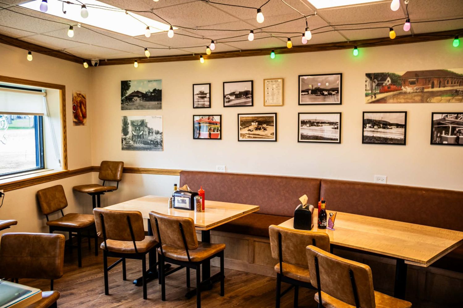 Cozy restaurant dining room with wooden tables, brown chairs, string lights, and black-and-white photos on cream walls.