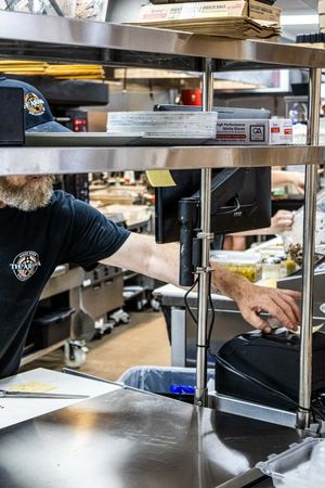 Bearded man in black t-shirt working in a busy commercial kitchen with stainless steel shelving and POS system.