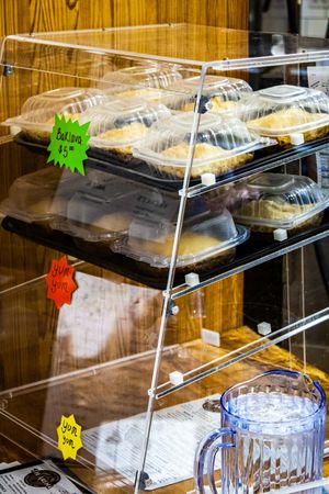 Bakery display case with packaged muffins and pastries on shelves, price tags visible, beside a blue glass pitcher.