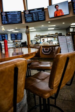 Interior of a casual restaurant with brown leather bar stools, wooden tables, menu boards, and a TV screen above the counter.