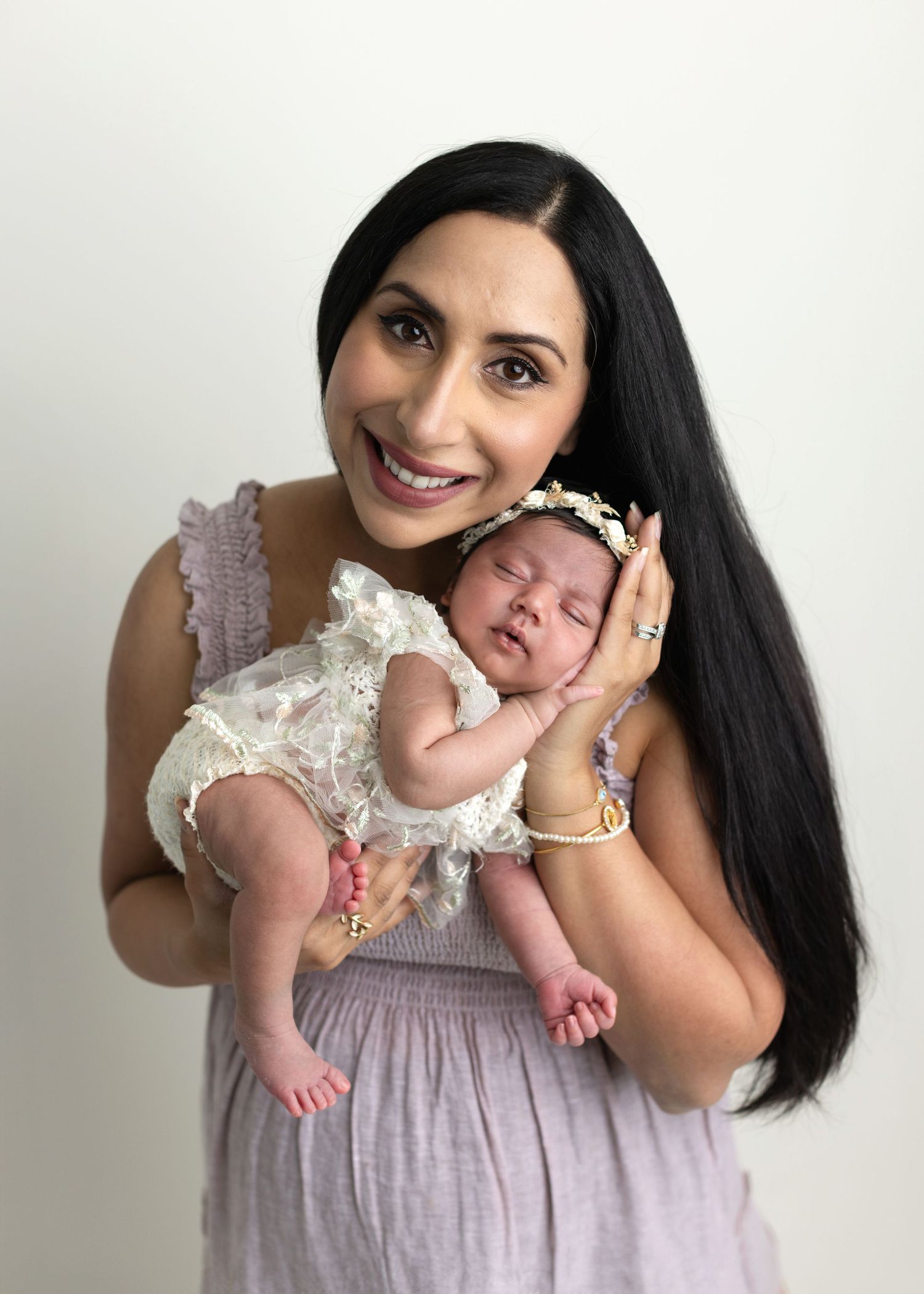 Smiling dark-haired woman in lavender dress holding a sleeping newborn baby girl wearing a floral headband.