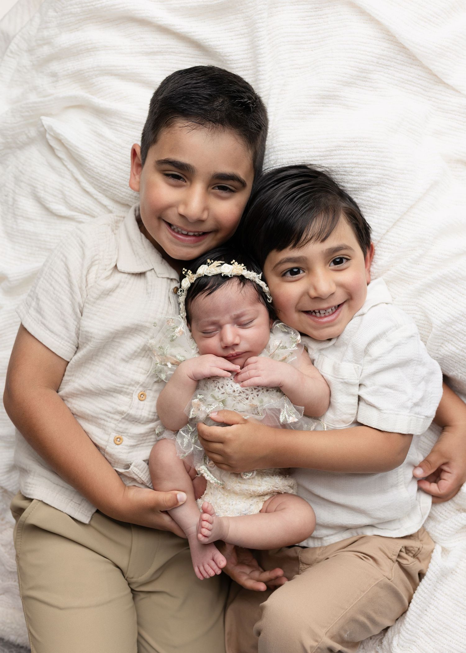 Two smiling boys pose with a sleeping newborn baby girl in a white floral headband on a white background.