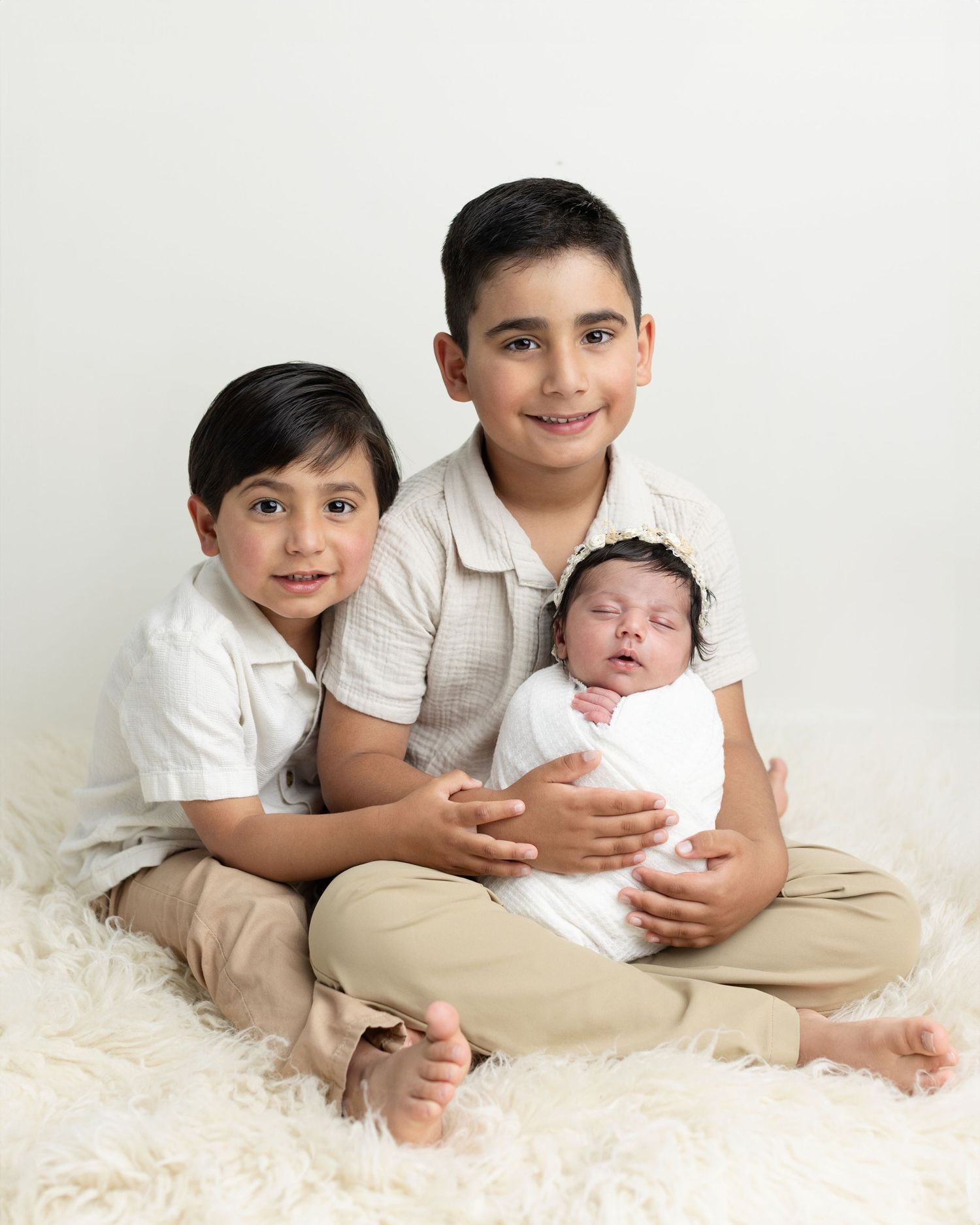 Two young brothers sitting cross-legged on a white rug, gently holding their swaddled newborn sibling in a studio portrait.
