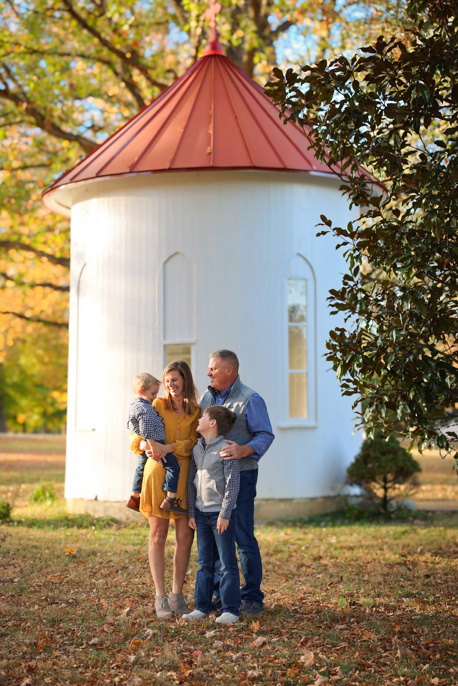 Happy family of four poses together near white silo surrounded by colorful autumn foliage.