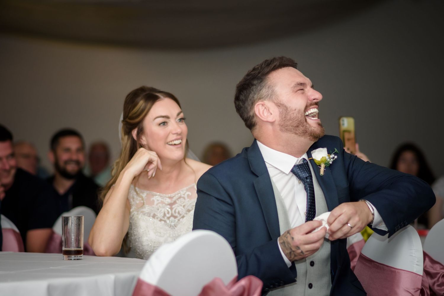 Bride and groom laughing joyfully at their wedding reception, seated at the head table in elegant attire.