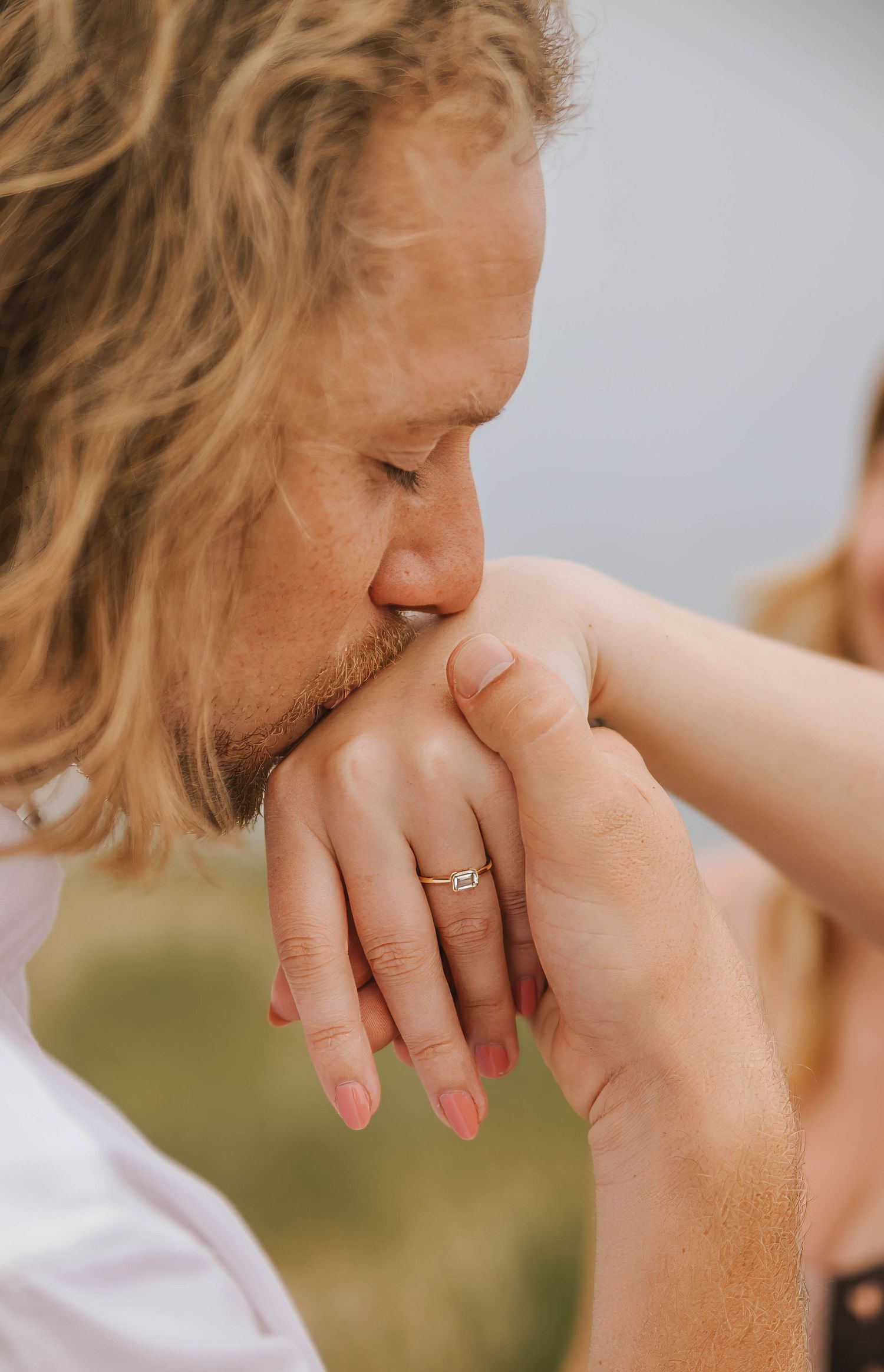 A man with curly blonde hair gently kisses a woman's hand adorned with a diamond engagement ring.