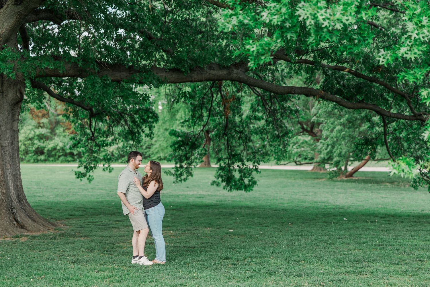 Couple embracing under a large tree in a lush green park during an outdoor engagement photo session.