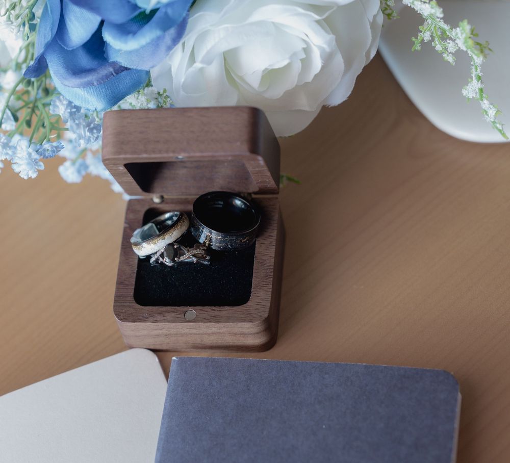 Wooden ring box holding two wedding bands on black velvet, beside white and blue flowers on a neutral surface.