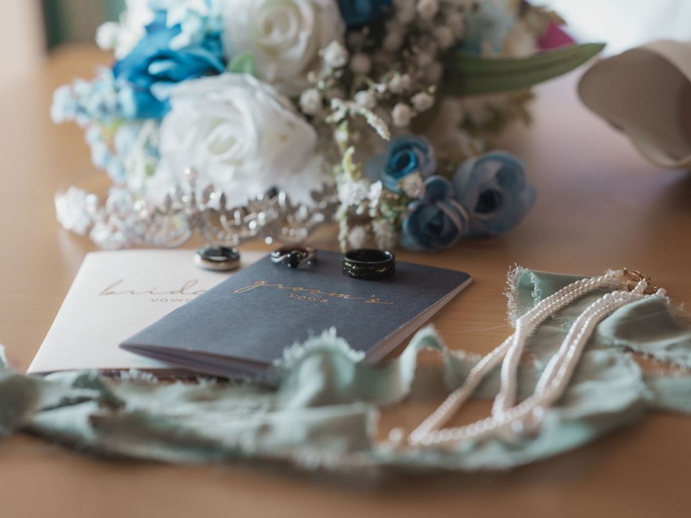 Wedding flat lay with rings, vows booklet, floral bouquet with blue and white flowers, and delicate accessories on a wooden surface.