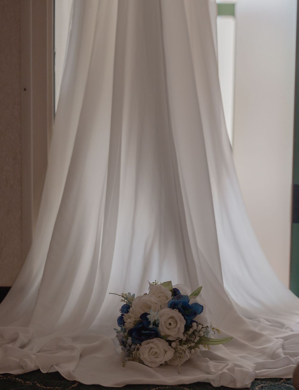White wedding dress train pooling on floor beside blue and white bridal bouquet with roses near a window.
