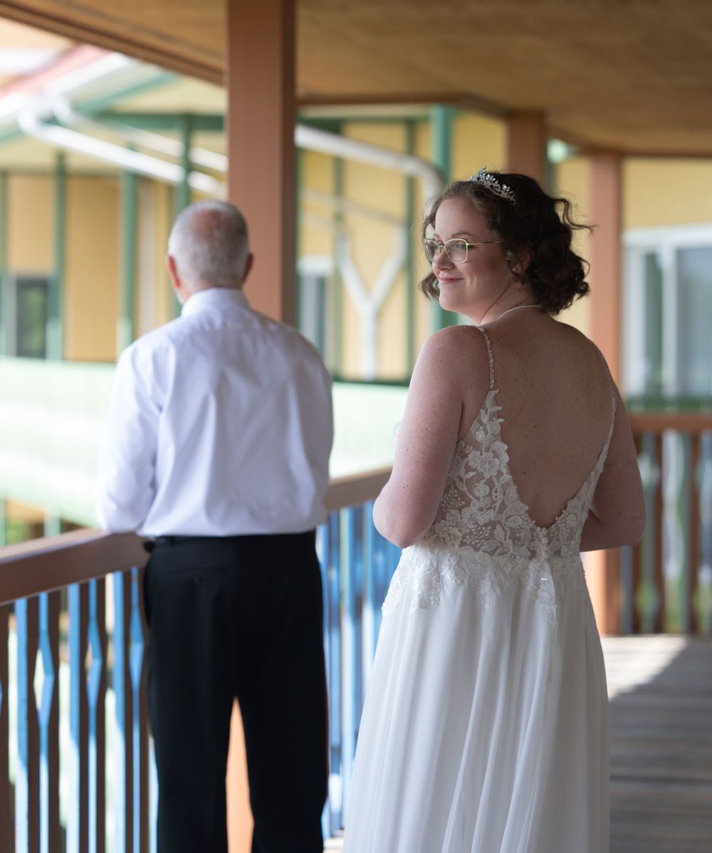 Bride in backless lace wedding gown smiles on wooden balcony as man in white shirt stands nearby.