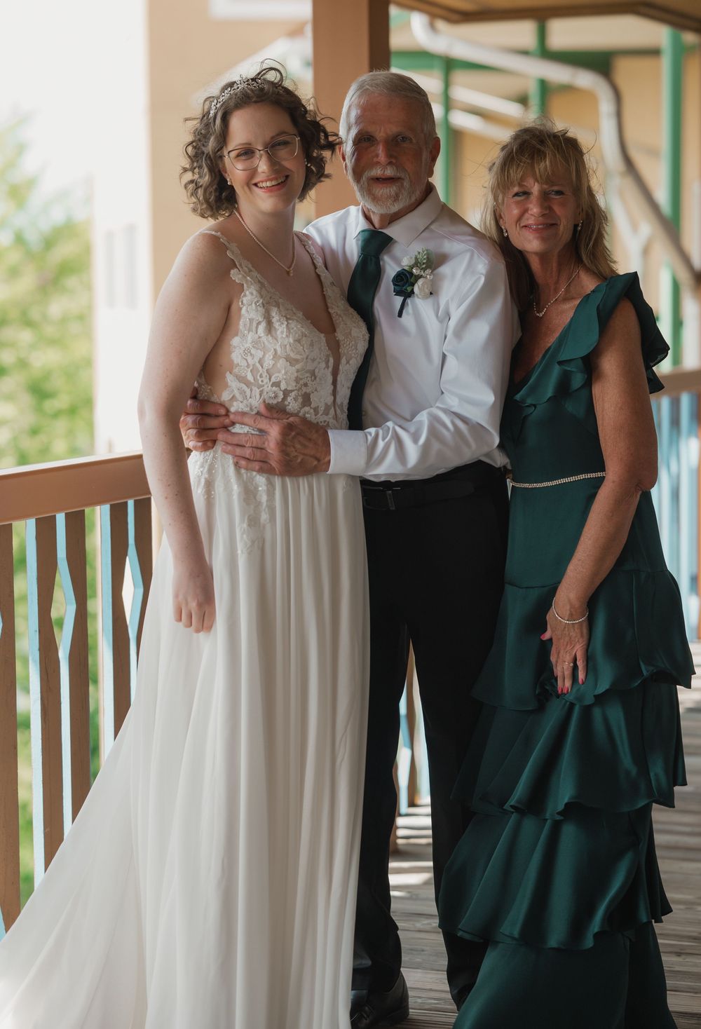 Bride in white lace gown poses with older couple in formal attire on wooden porch at wedding.