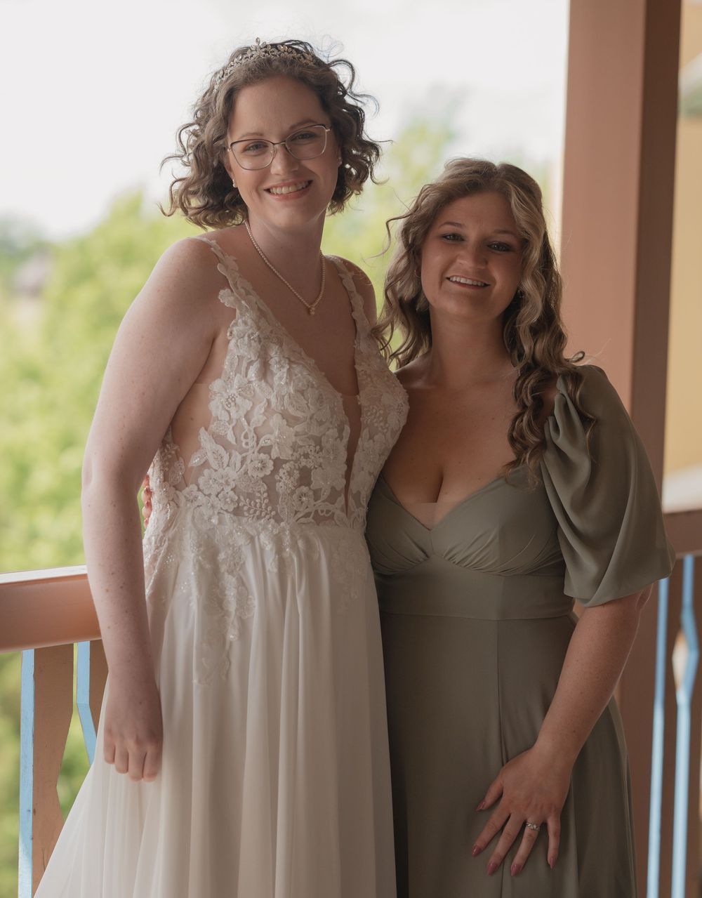 Bride in lace wedding gown poses with bridesmaid in sage green dress on outdoor balcony.