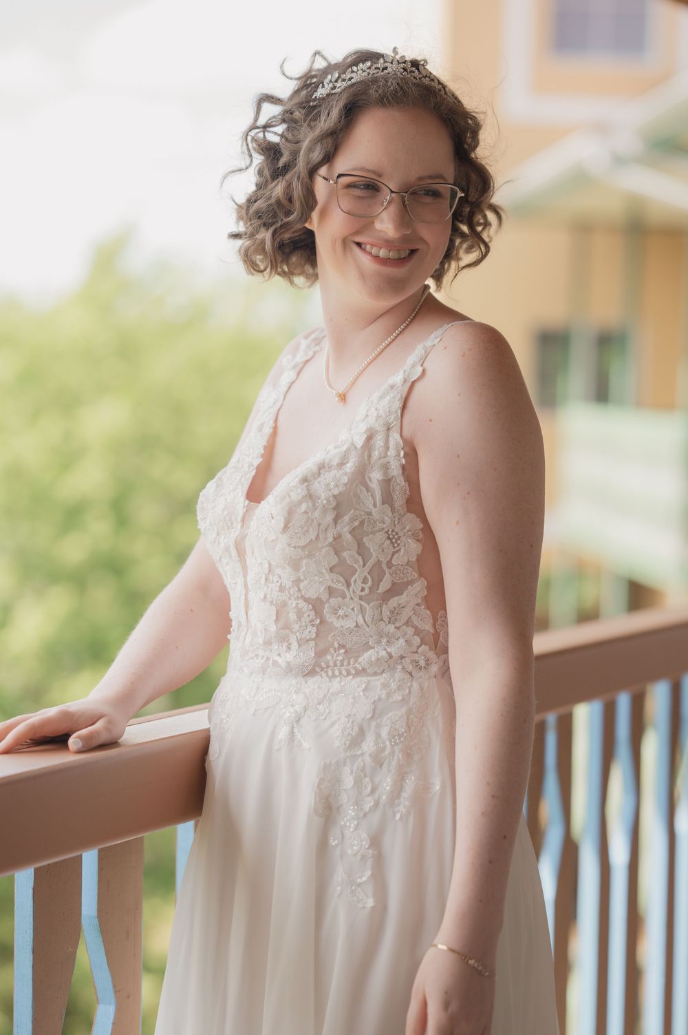 Smiling bride in lace wedding gown and tiara poses on balcony railing outdoors.