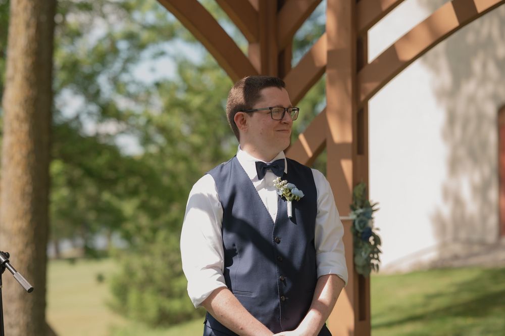 Groom in navy vest and bow tie with boutonniere stands under wooden pergola at outdoor wedding ceremony.