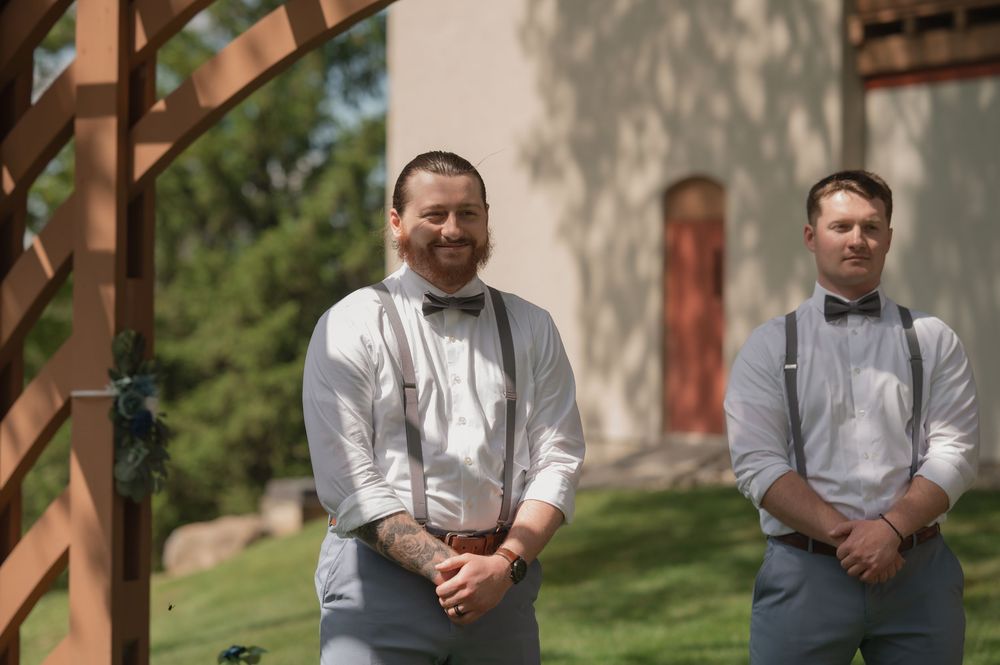 Groom and groomsman in gray suspenders and bow ties stand outdoors near wooden arbor at wedding.