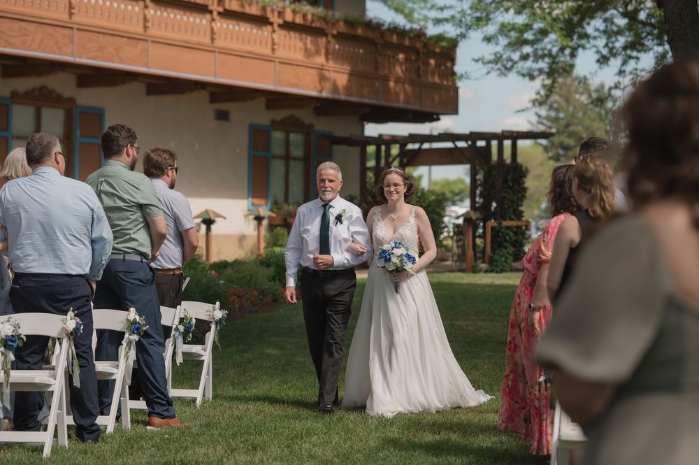 Bride walks down outdoor aisle with escort at elegant garden wedding ceremony, guests seated in white chairs.