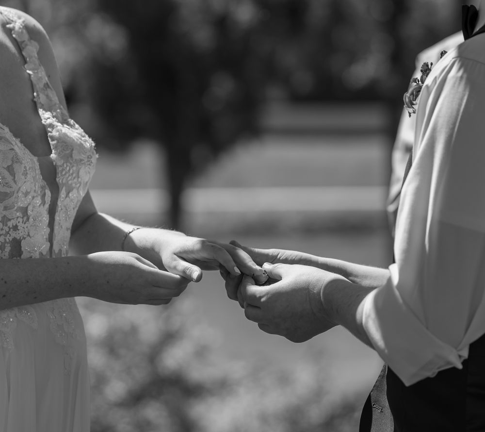 Black and white photo of a bride and groom exchanging rings during their wedding ceremony outdoors.