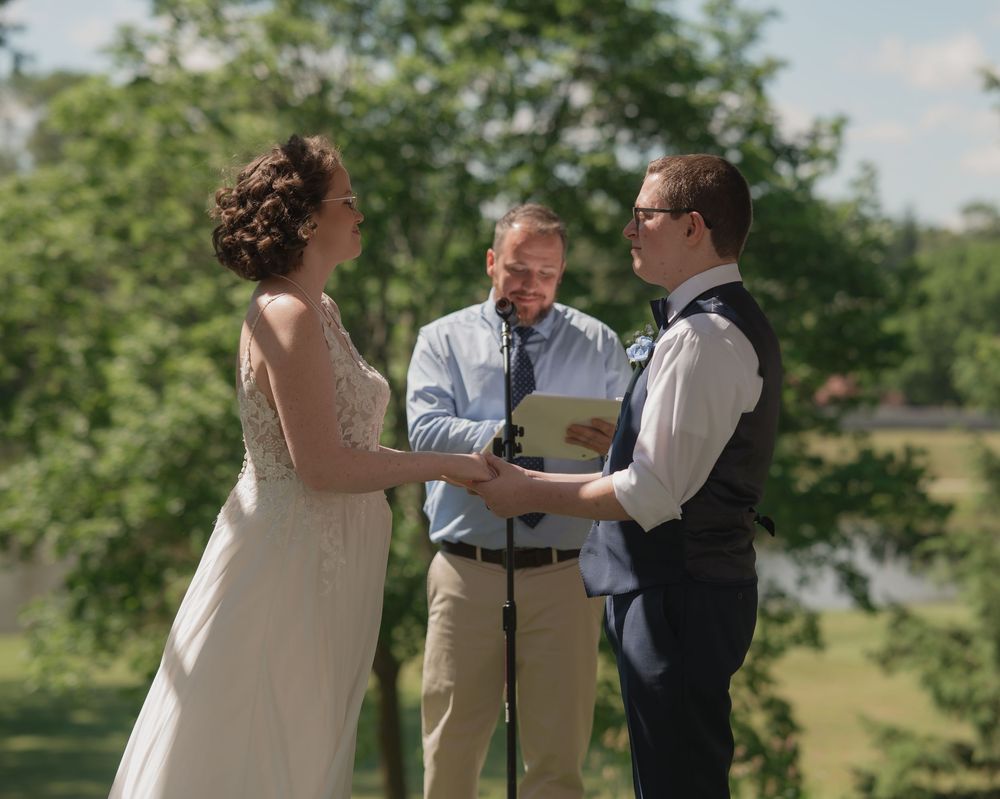 Outdoor wedding ceremony with bride in white lace gown, groom in vest, and officiant reading vows in lush green setting.