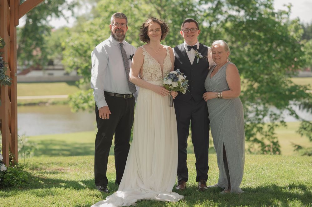 Wedding party of four posing outdoors on green grass with trees and a pond in the background on a sunny day.