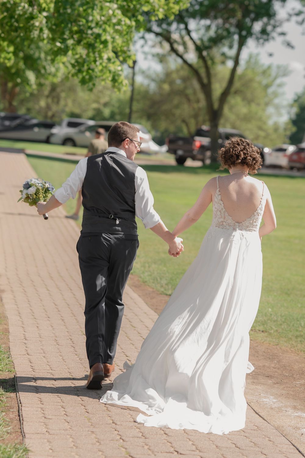 Bride and groom walking hand in hand on brick path, bride in backless white gown, groom in black suit with gray vest.