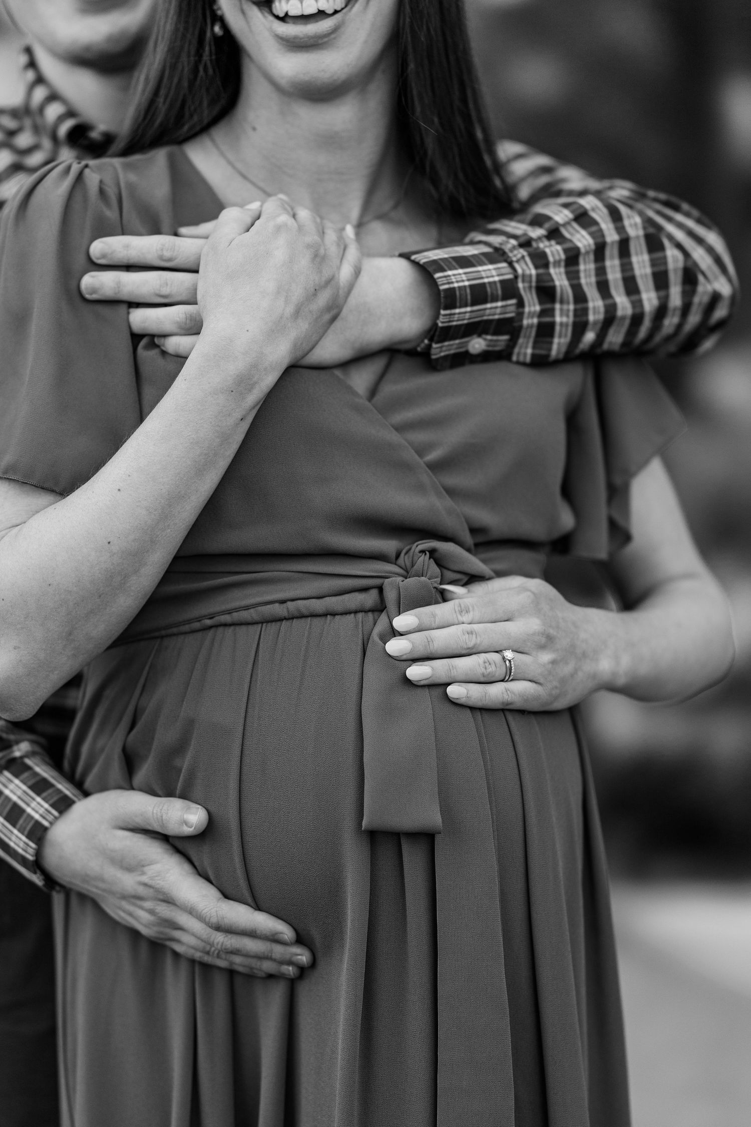 Black and white photo of a smiling pregnant woman being embraced by her partner, both touching her baby bump.