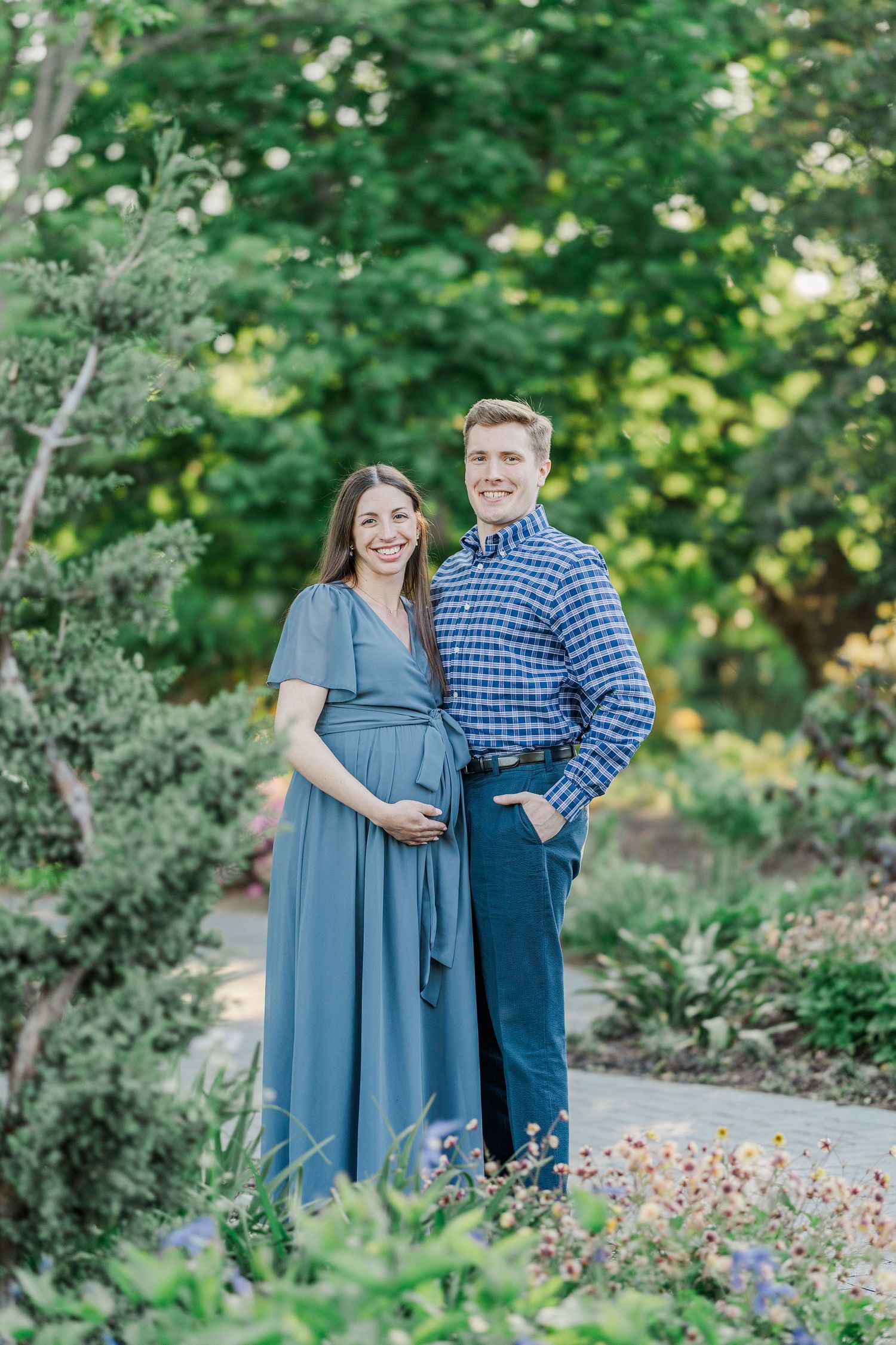 Smiling pregnant couple poses together in a lush garden, woman in blue maternity dress, man in plaid shirt.