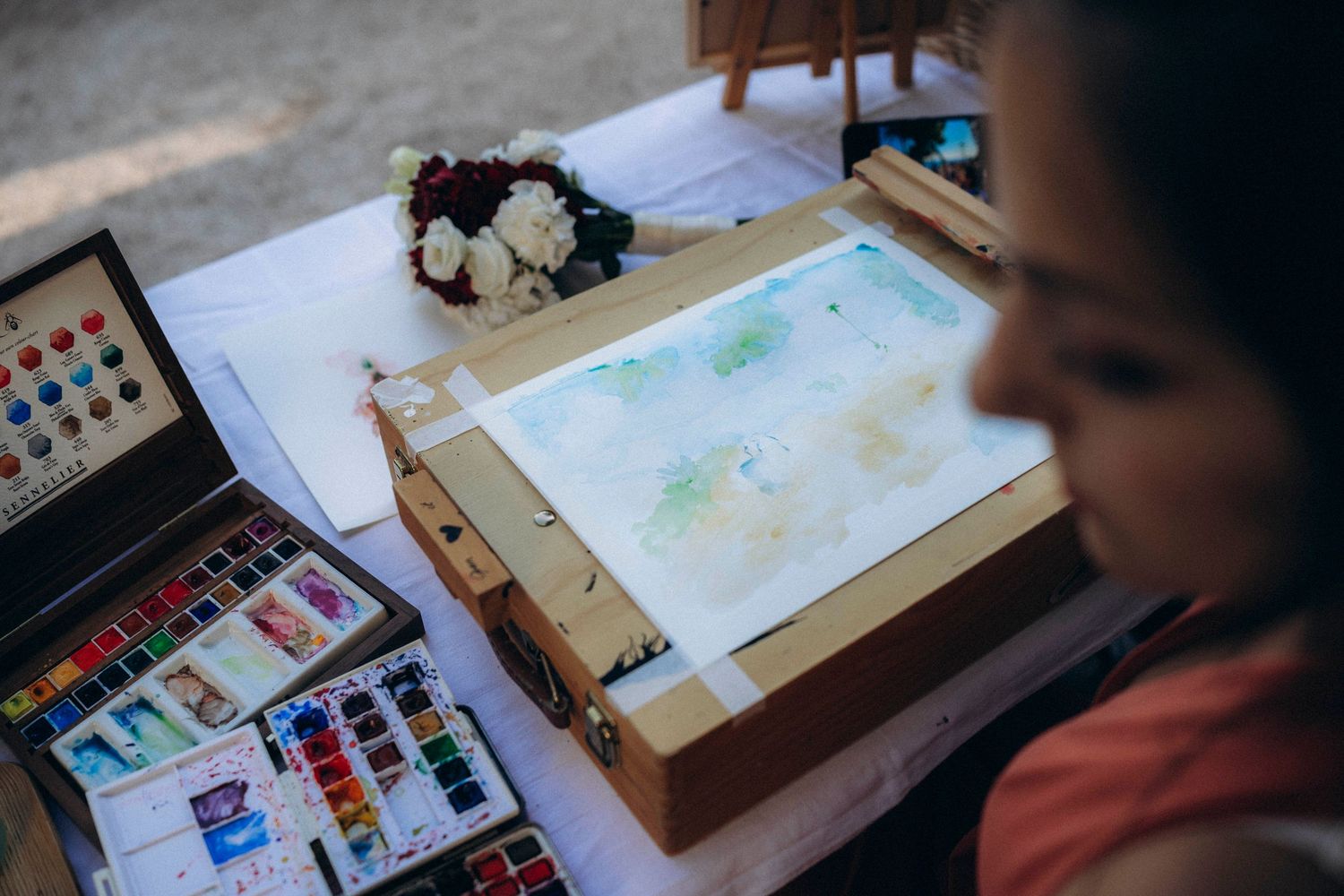 Artist painting watercolors on a light box outdoors, with colorful paint palettes and flowers nearby.