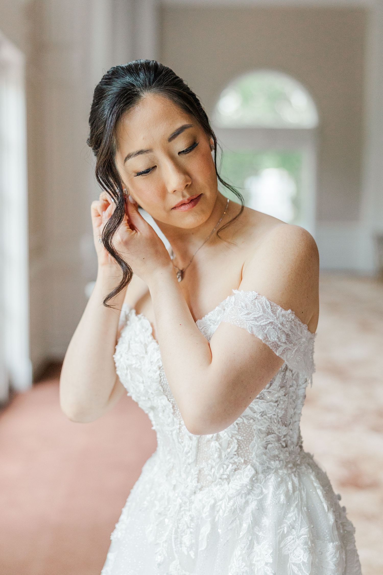 Asian bride in off-shoulder white lace wedding gown adjusting hair, elegant bridal portrait in soft natural light.