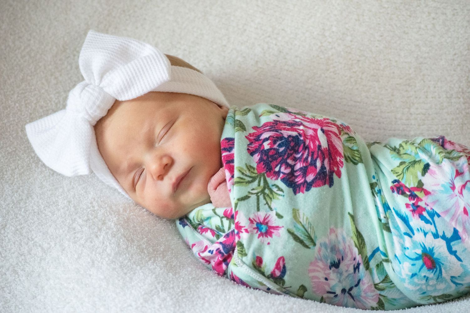 Sleeping newborn baby swaddled in floral blanket wearing white bow headband.