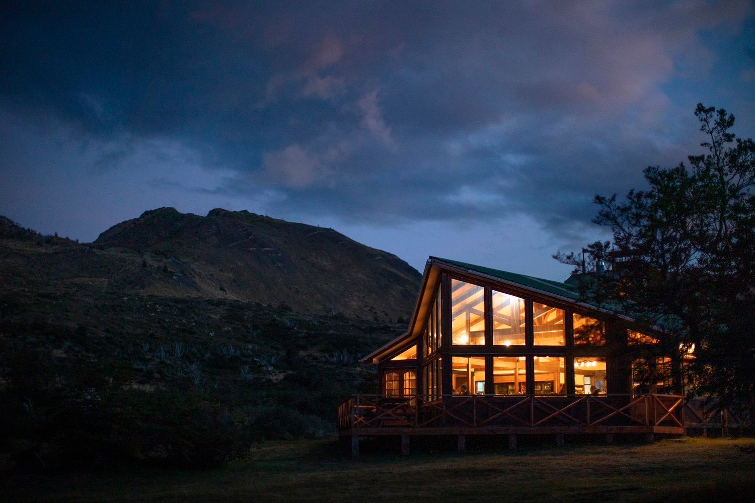 Rustic log cabin glowing warmly at dusk, surrounded by tall pine trees against a dramatic twilight sky.