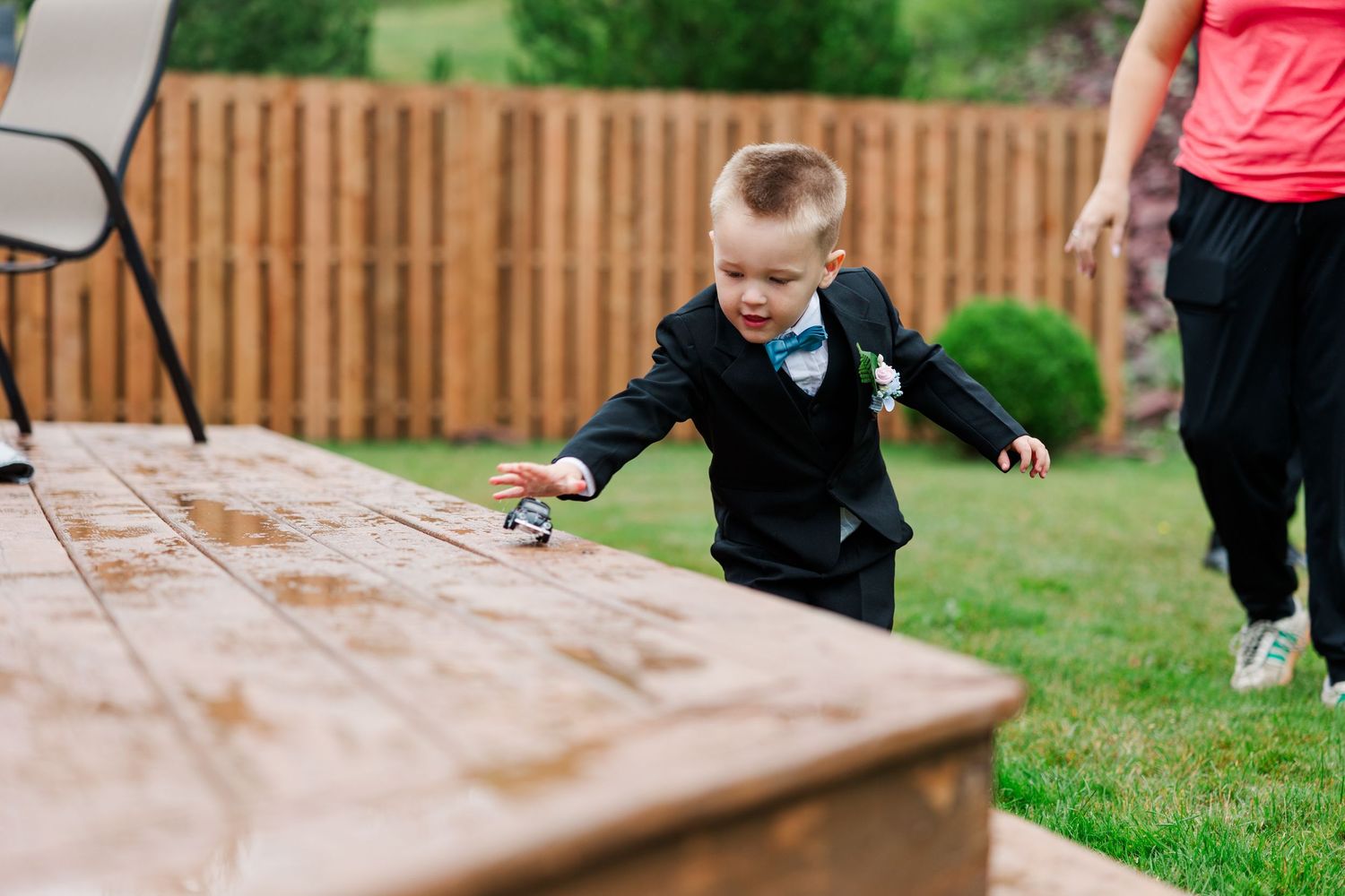 Young boy in black suit with bow tie playing at wooden table outdoors at a backyard wedding celebration.
