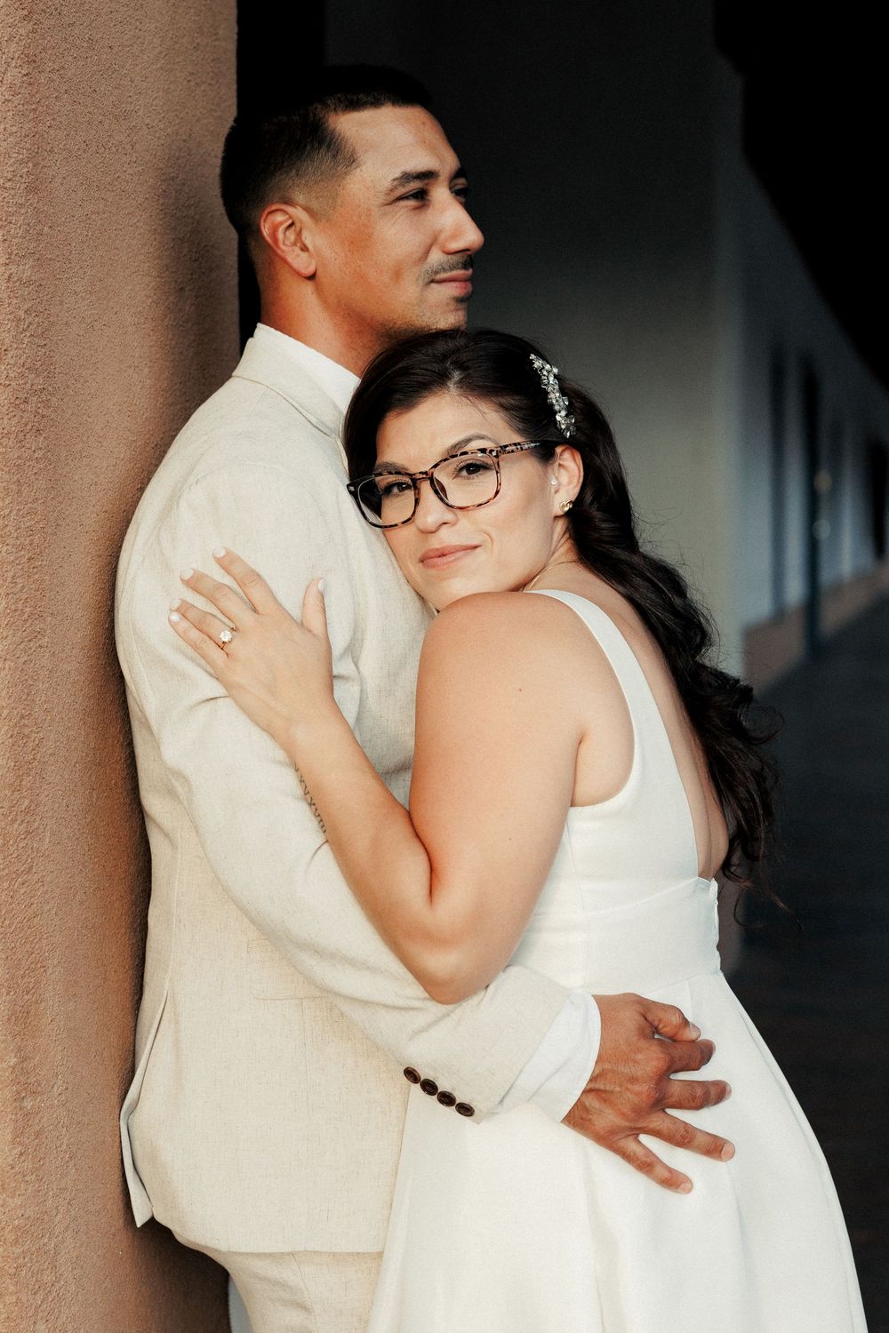 Bride in white dress and glasses embraces groom in beige suit against a column in a romantic wedding portrait.