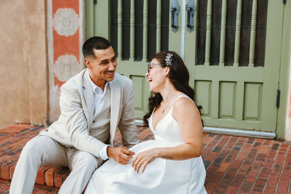 A joyful bride and groom sit on brick steps, laughing and holding hands, dressed in white gown and light gray suit.
