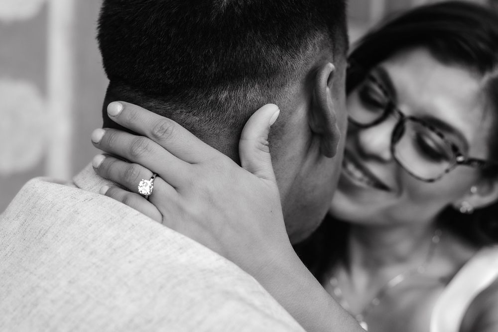 Black and white photo of a smiling woman with glasses placing her hand on a mans neck showing off her engagement ring.