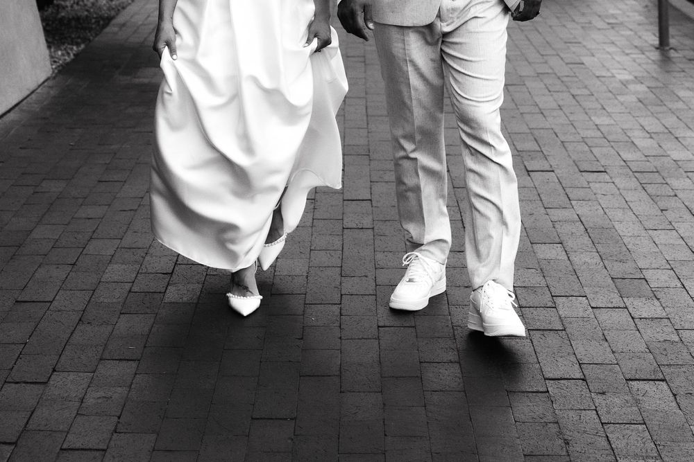 Black and white photo of a bride in white dress and groom in light suit walking on brick pavement.
