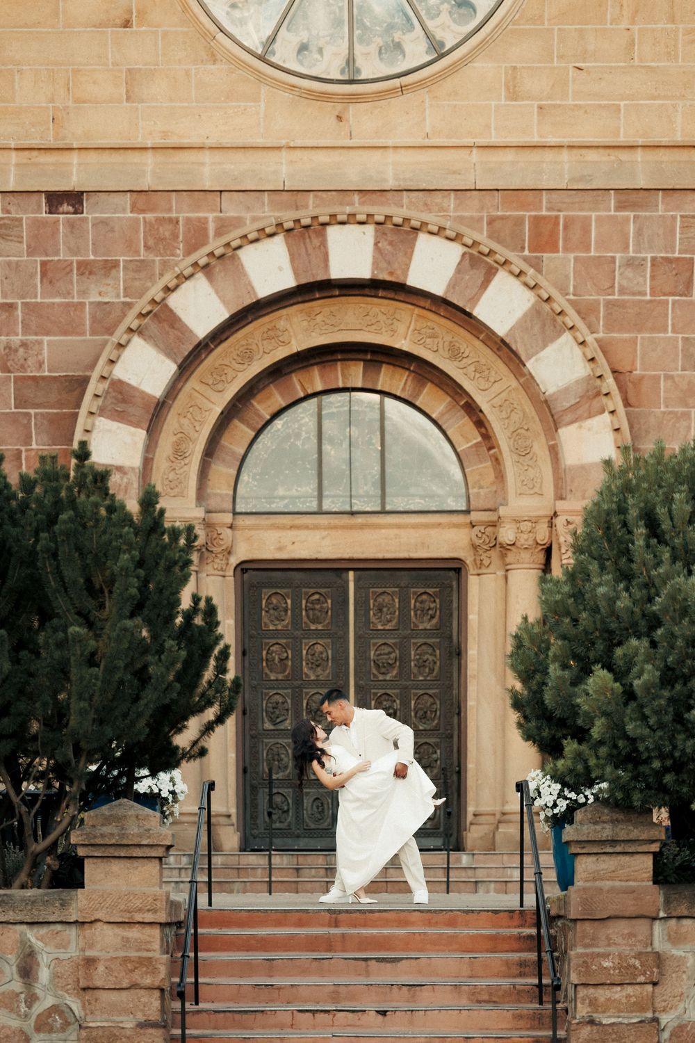 Couple sharing a romantic dip kiss on church steps, bride in white gown, ornate arched doorway and stone facade backdrop.