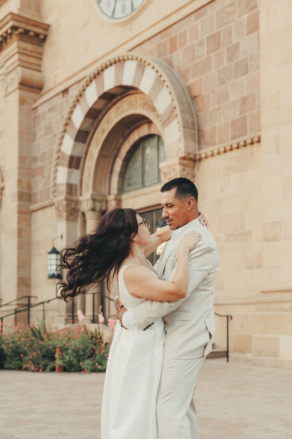 Couple embracing and dancing in front of a historic stone church building, woman in white dress with hair blowing in wind.