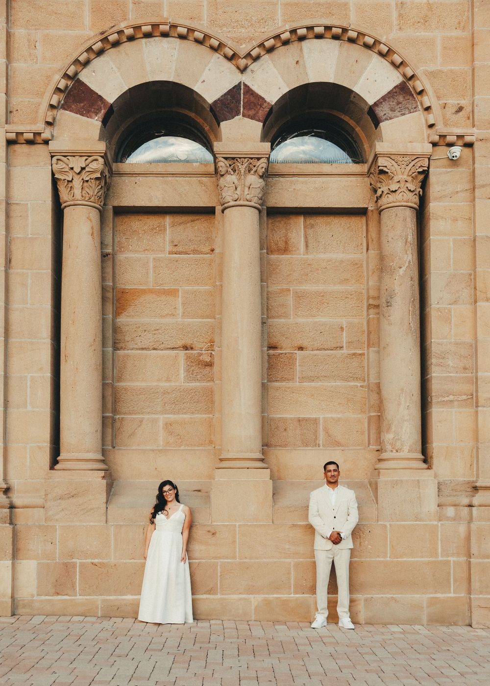 Bride in white gown and groom in cream suit pose apart before ornate stone arched windows.