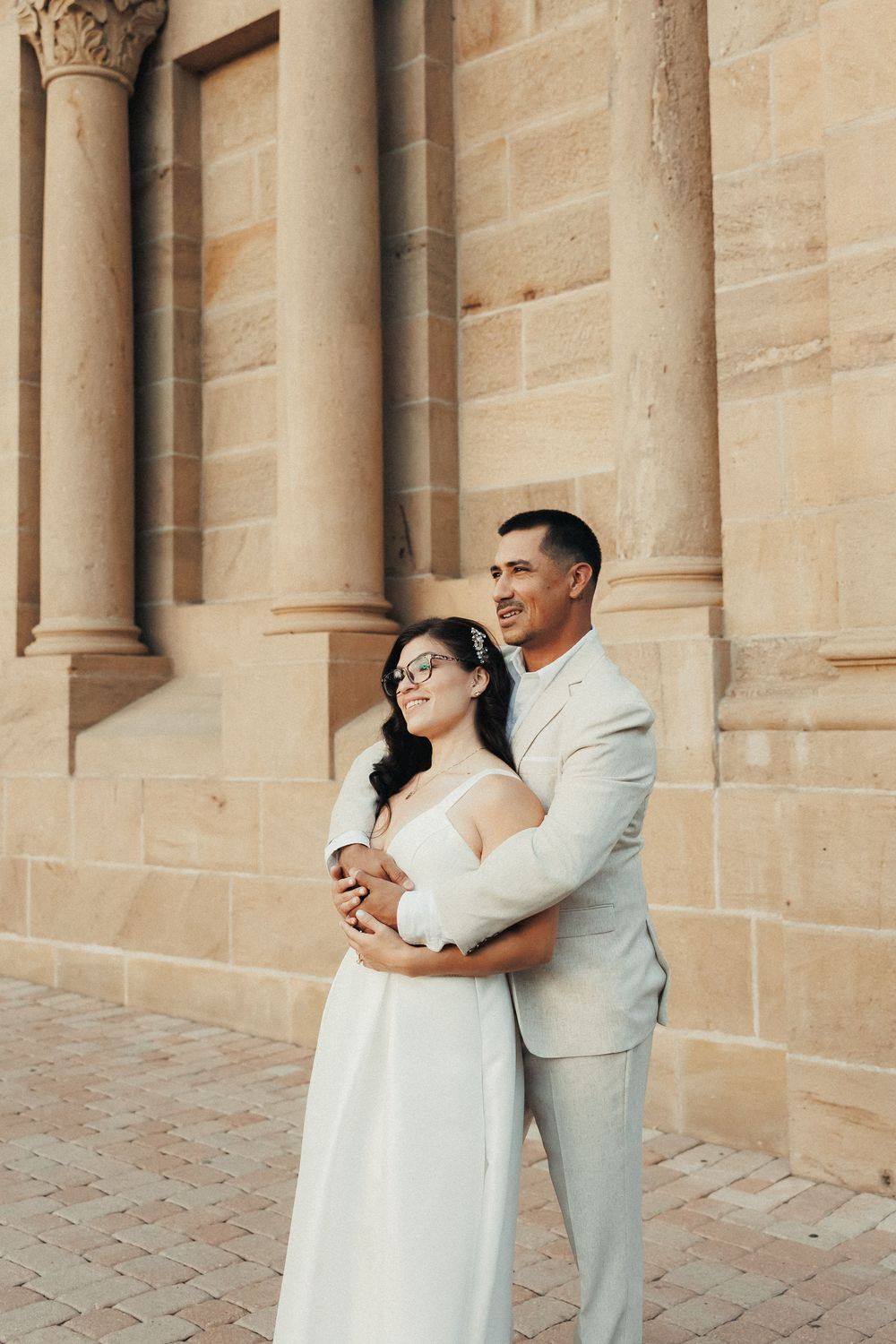 Bride and groom embracing in front of a sandstone building, wearing white dress and beige suit.
