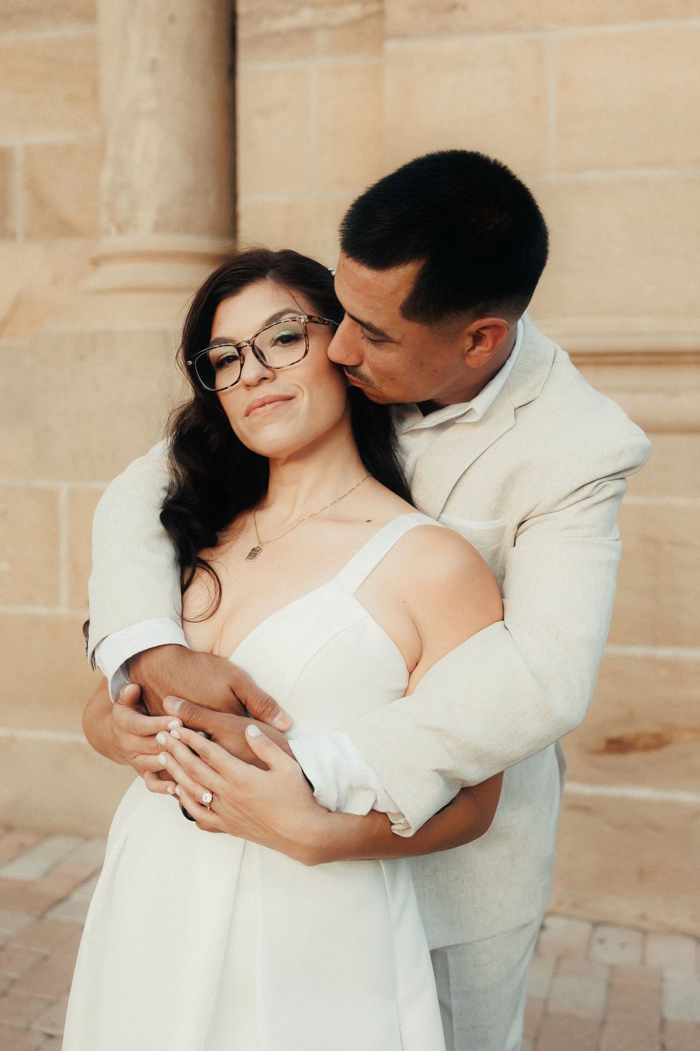 A groom in a beige suit embraces his bride in a white dress with glasses, sharing a tender kiss outdoors.