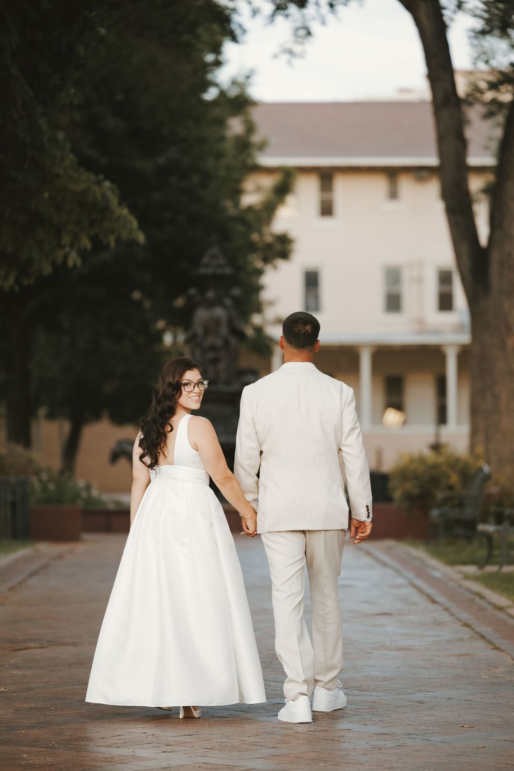Bride in white gown and groom in beige suit walk hand-in-hand toward a white colonial venue at dusk.