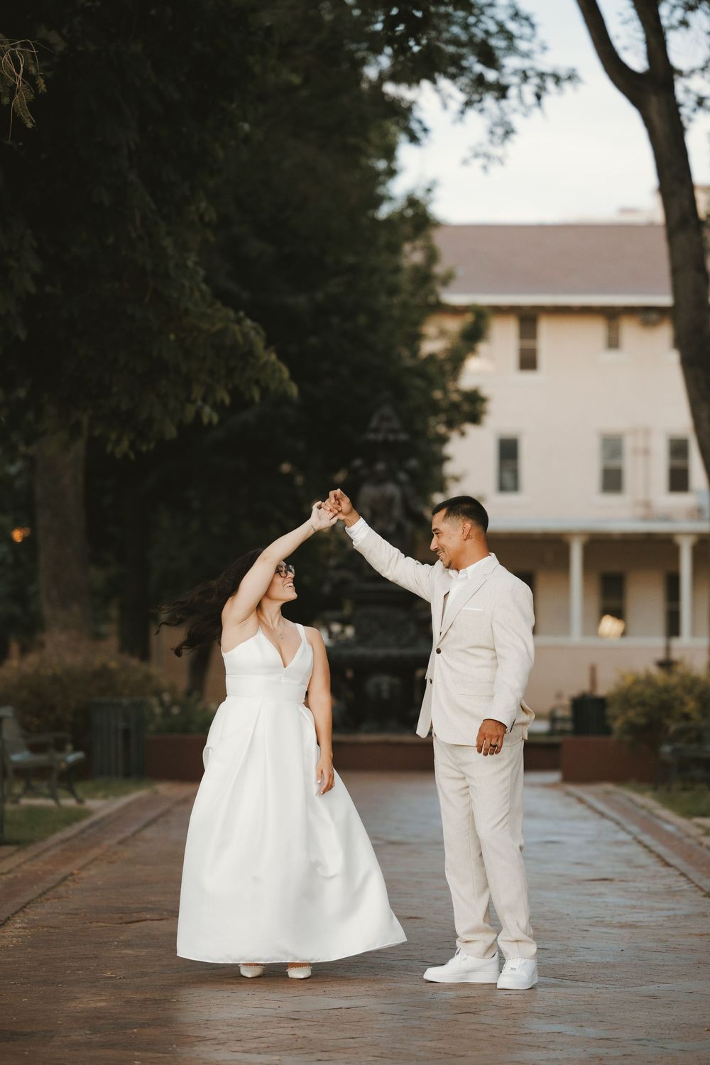 Bride and groom dancing outdoors on a brick path, with a white historic building and lush trees in the background.